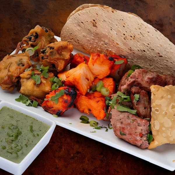 A white platter of various Indian appetizers, including pakoras, tandoori chicken, and seekh kebab, with green chutney.